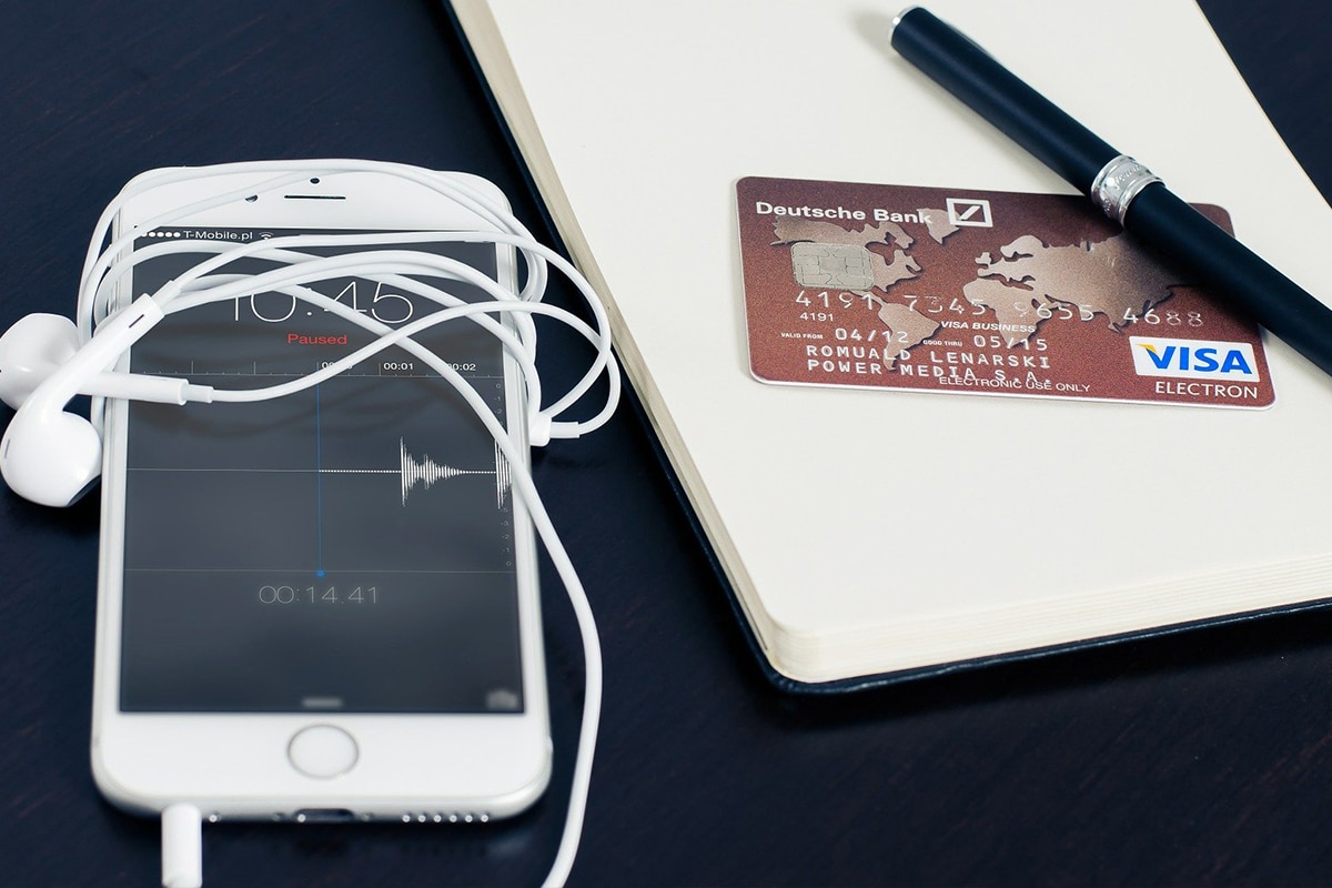iPhone com fone de ouvido enrolado em volta, um bloco de notas, uma caneta preta e um cartão de crédito