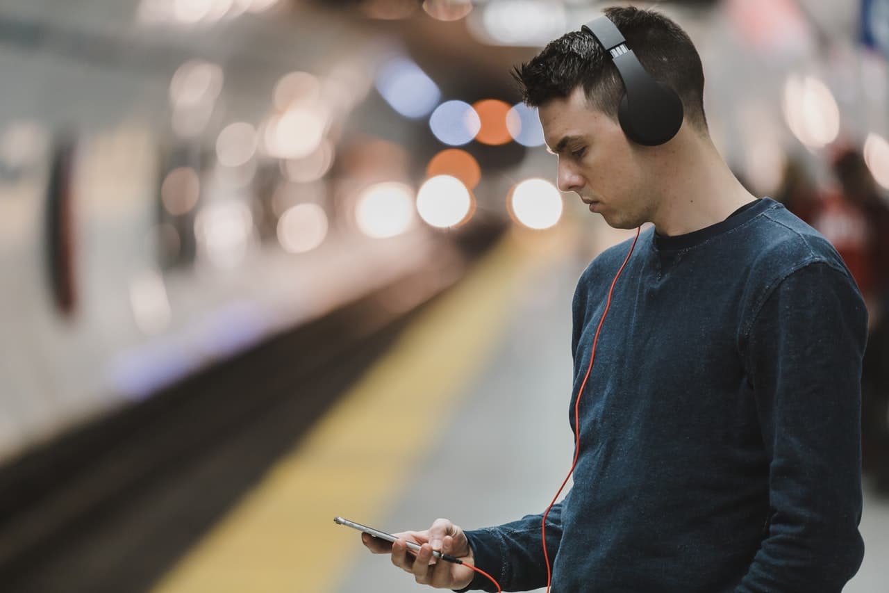 homem na plataforma do metrô, mexendo no celular e com fones de ouvido tipo headset