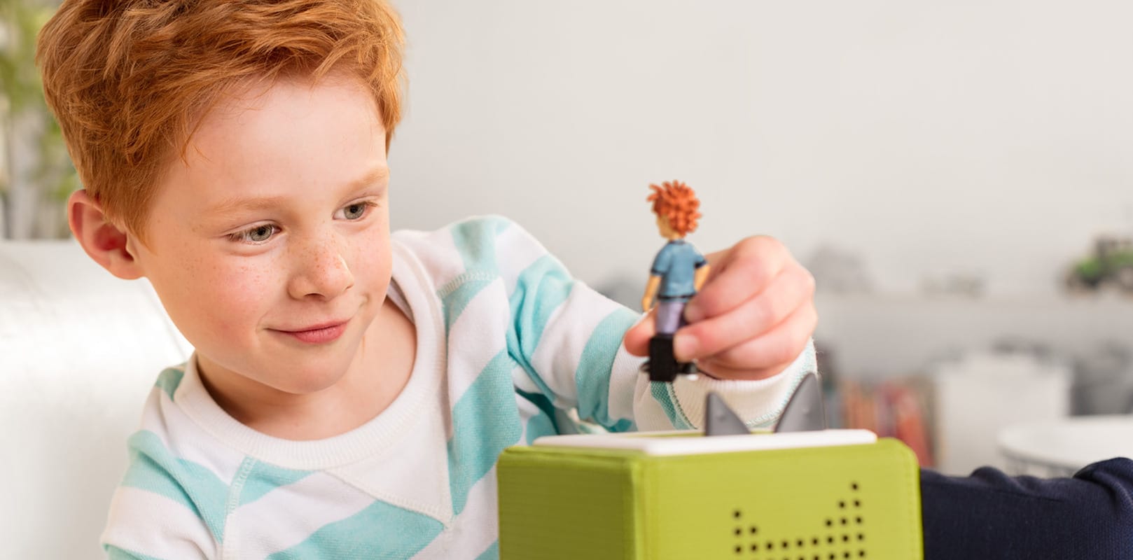 Caixas de som tonies utiliza bonecos RFID - imagem mostra um menino ruivo deitado na sala de casa brincando com um tonie, boneco ruivo em cima de uma caixa de som verde - tecnologia é a mesma utilizada nos amiibos