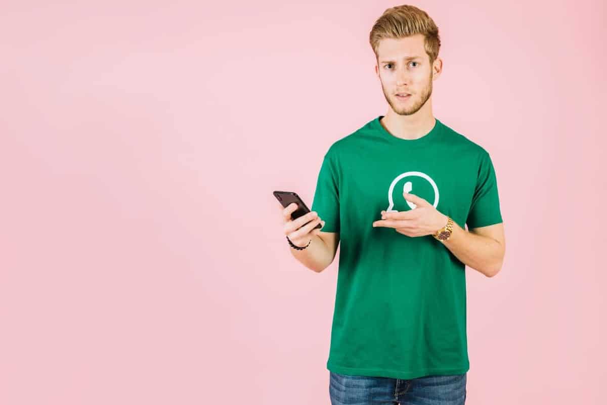 homem usando camiseta com símbolo do WhatsApp segura celular com cara de dúvida