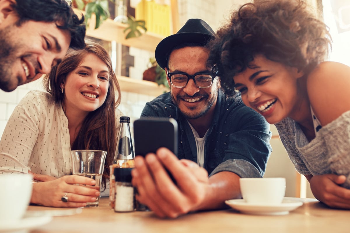 Foto de amigos reunidos em uma mesa e usando o celular para mandar áudio em rede social