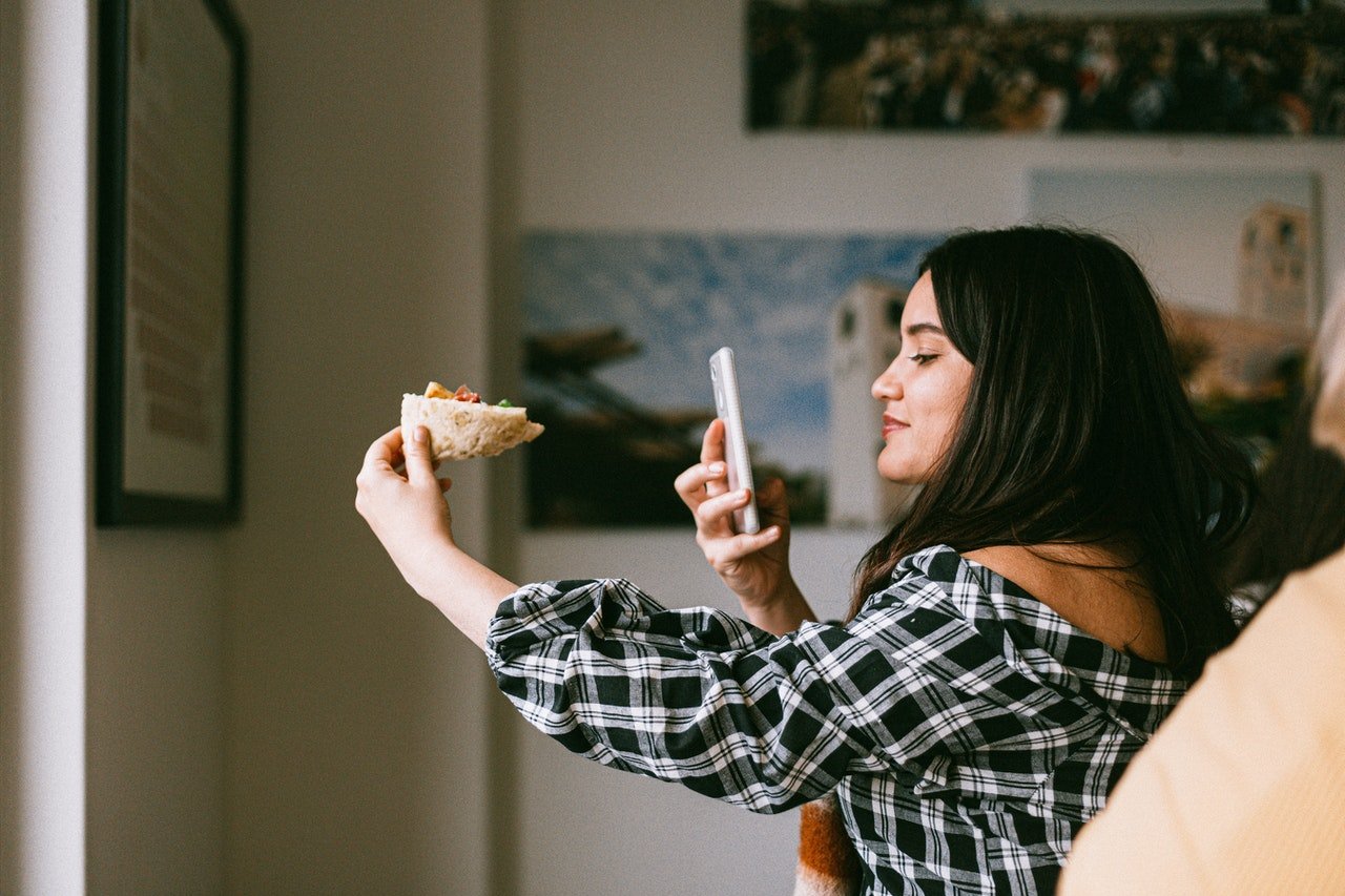 moça apontando celular para comida que está segurando com uma das mãos para fazer foto ou vídeo