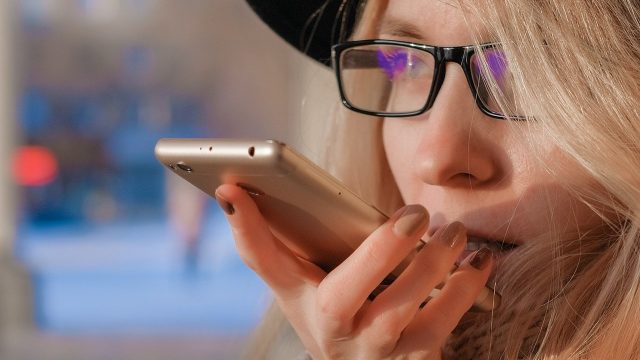 Mulher loira de óculos e chapéu mandando áudio no celular
