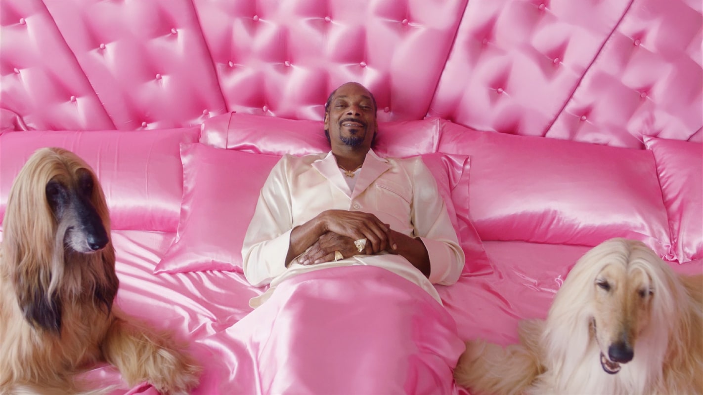 Snoop Dogg em uma cama cor de rosa com um cachorro em cada lado deitado com ele