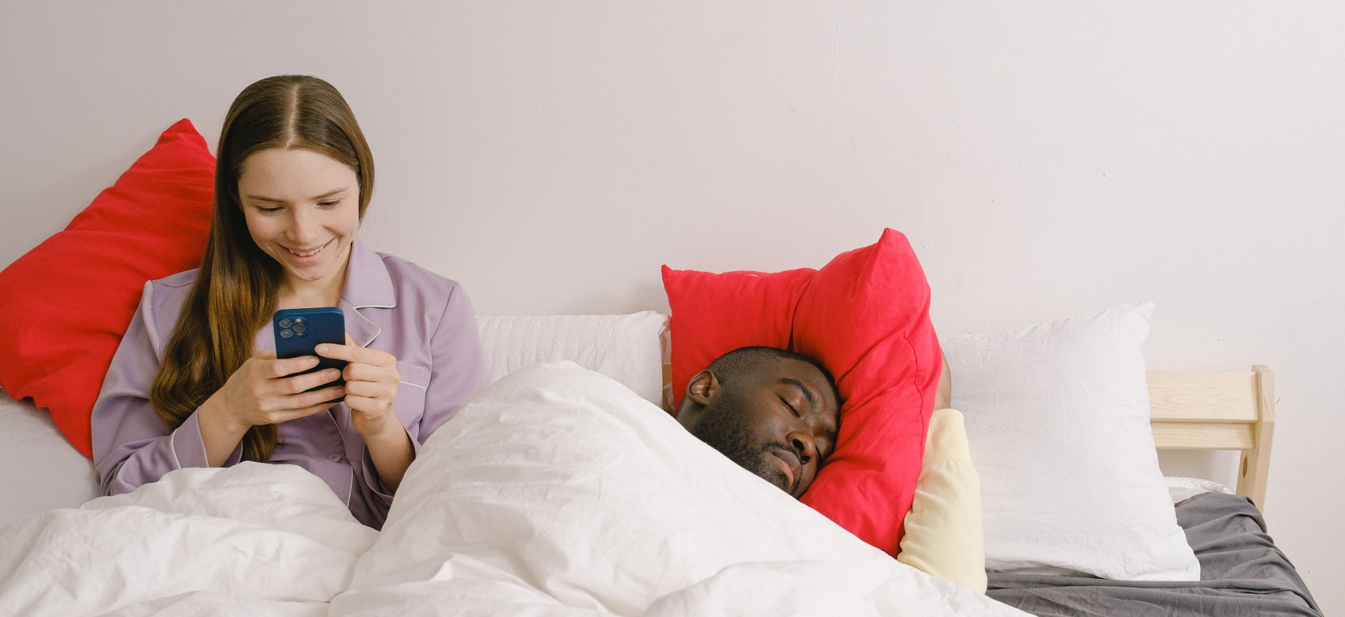 Imagem mostra uma mulher mexendo no celular sentada na cama e um homem ao lado dela, dormindo. Amazon conseguiu liberação para monitorar o sono dos usuários por meio de radar