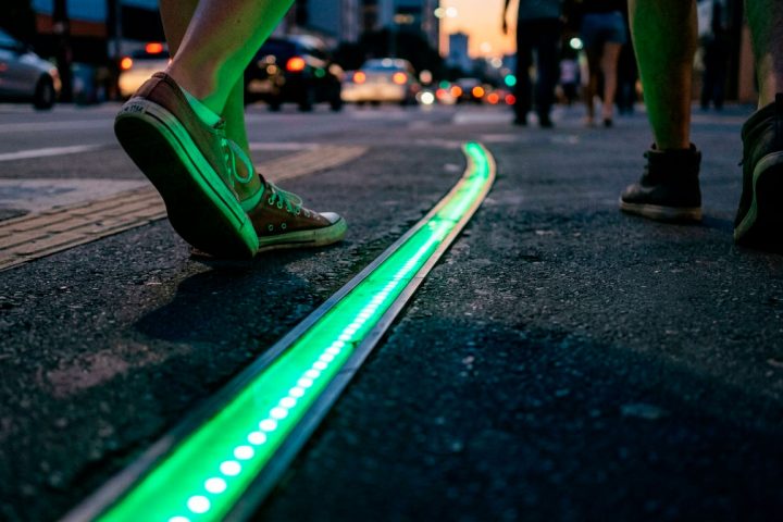 Luzes de LED embutidas na calçada indicando o sinal de trânsito, com pés de pedestres ao lado.